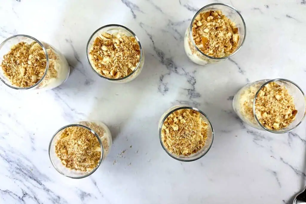 Six glasses filled with a dessert topped with crushed nuts or crumbs are arranged on a white marble surface.