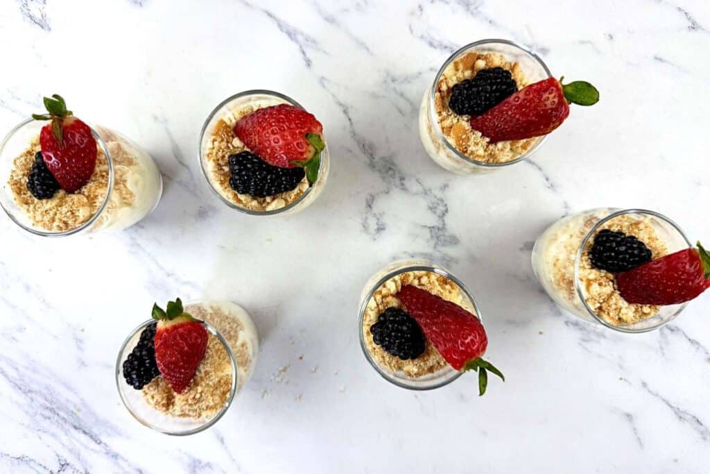 Six small dessert cups topped with crumbled crumbs, a strawberry, and a blackberry are arranged on a white marble surface.