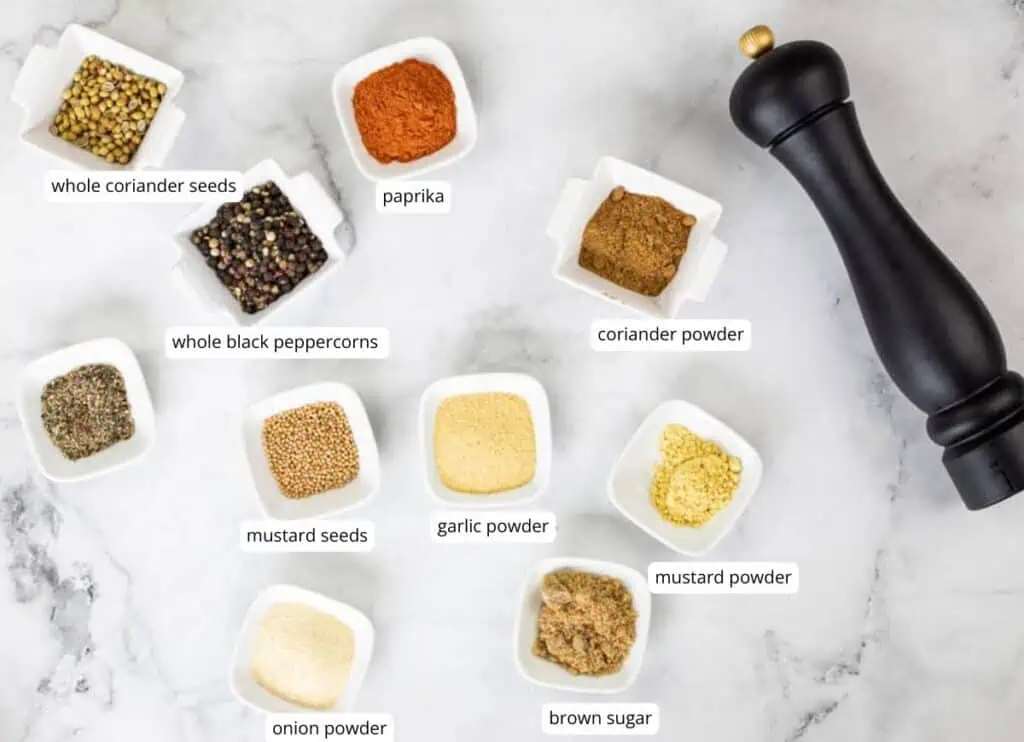 Assorted spices and seasonings in small white dishes, labeled with names, arranged next to a black pepper grinder on a white marble surface.