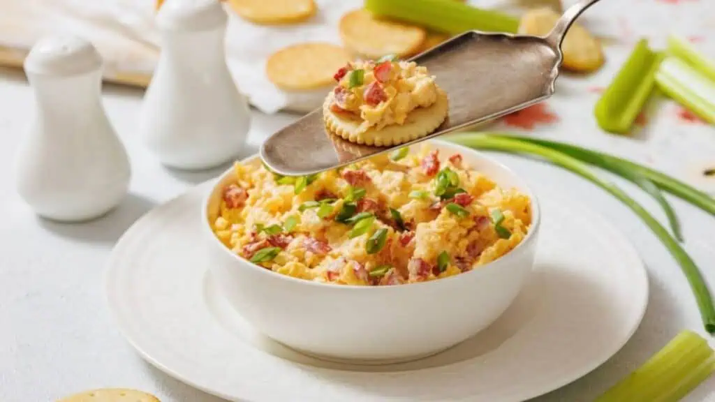 A bowl of pimento cheese dip garnished with green onions, with a cracker topped with the dip held above it on a serving utensil. Crackers and celery sticks are nearby.