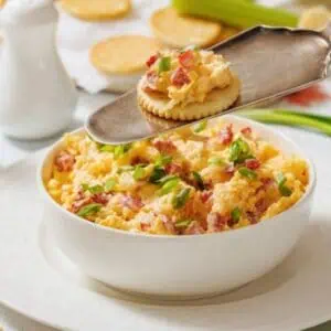 A bowl of pimento cheese dip garnished with green onions, with a cracker topped with the dip held above it on a serving utensil. Crackers and celery sticks are nearby.