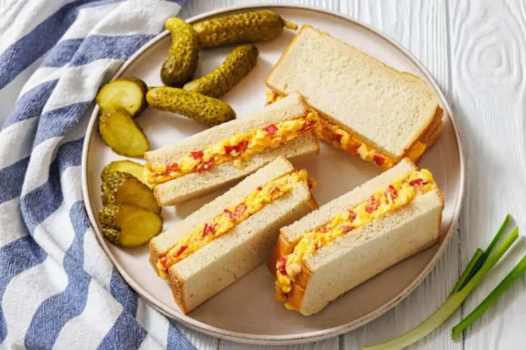 A plate with pimento cheese sandwiches cut into halves, served with whole and sliced pickles, on a white table with a striped cloth napkin.