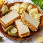 A plate with three halves of pimento cheese sandwiches, potato chips, and pickles, with lettuce and more chips scattered on the marble surface.