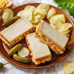 A plate with three halves of pimento cheese sandwiches, potato chips, and pickles, with lettuce and more chips scattered on the marble surface.