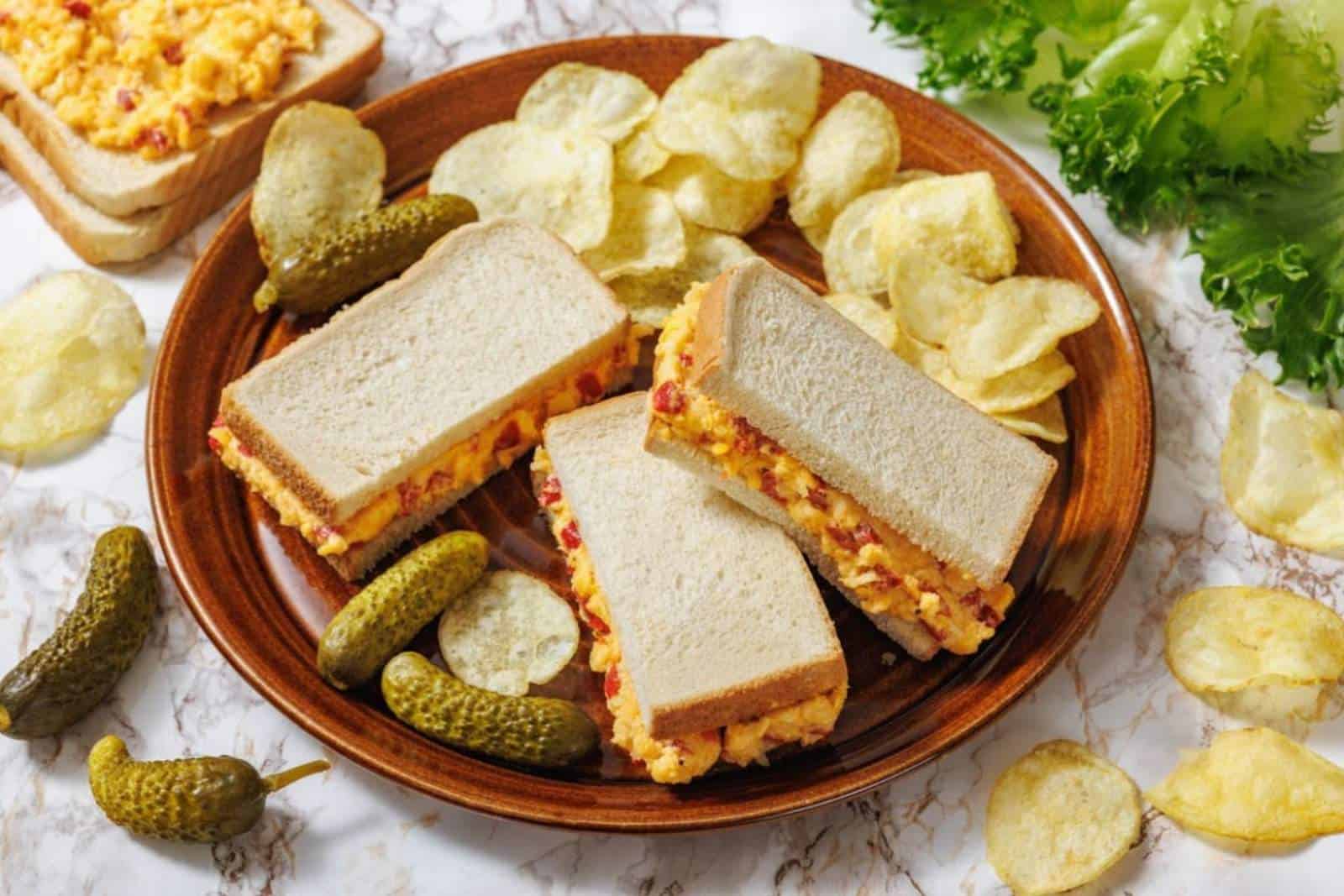 A plate with three halves of pimento cheese sandwiches, potato chips, and pickles, with lettuce and more chips scattered on the marble surface.