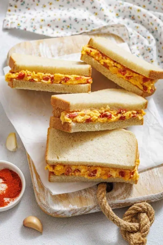 Four sandwiches filled with scrambled eggs and red peppers are stacked on a wooden board lined with parchment paper; a bowl of paprika and garlic cloves are nearby.