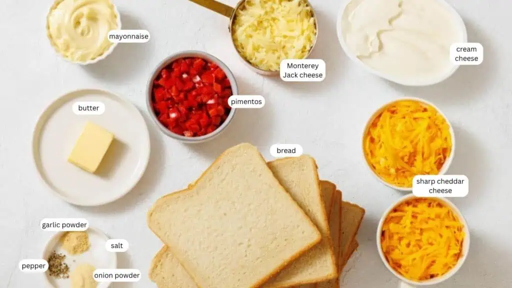 Slices of bread, shredded Monterey Jack cheese, sharp cheddar cheese, cream cheese, mayonnaise, pimentos, butter, salt, pepper, garlic powder, and onion powder arranged on a table.
