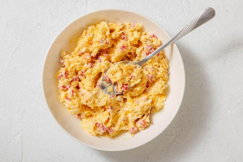 A bowl of pimento cheese spread with a spoon, featuring a mixture of shredded cheese, mayonnaise, and diced pimentos.