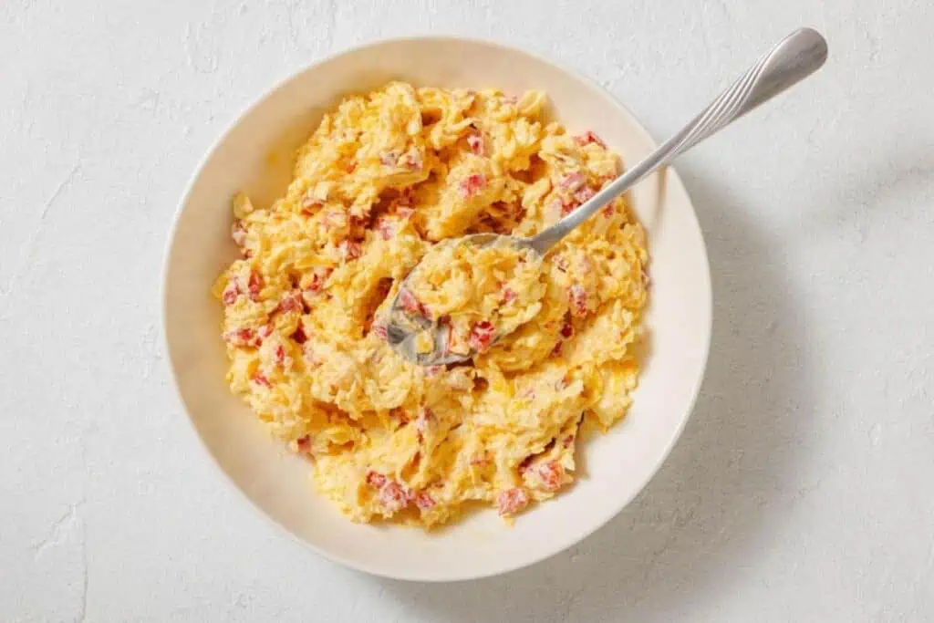 A bowl of pimento cheese spread with a spoon, featuring a mixture of shredded cheese, mayonnaise, and diced pimentos.