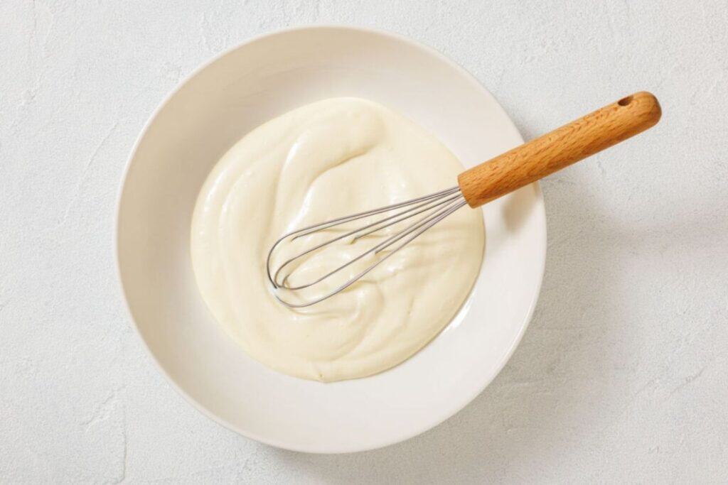 A metal whisk rests in a white bowl filled with a smooth, creamy white mixture on a light-colored surface.