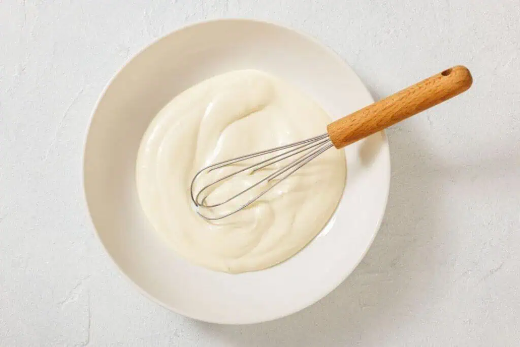 A metal whisk rests in a white bowl filled with a smooth, creamy white mixture on a light-colored surface.