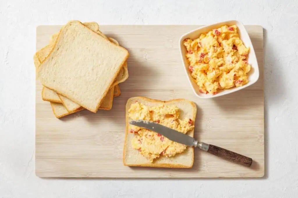A stack of sliced white bread next to a bowl of pimento cheese spread, with one slice being spread on a cutting board using a knife.