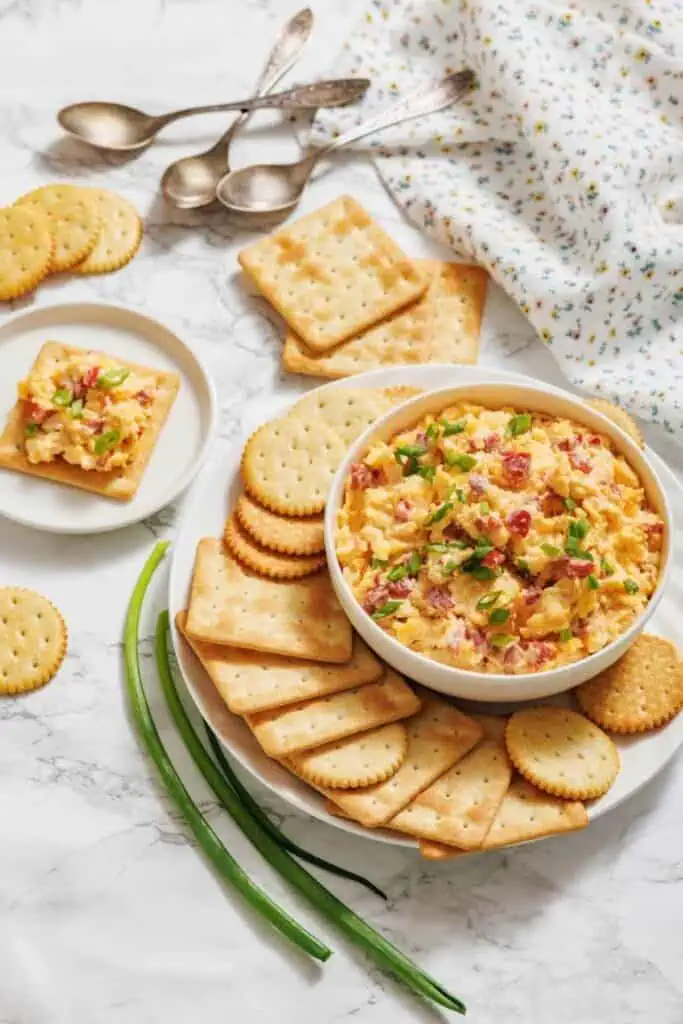 A bowl of pimento cheese dip is surrounded by various crackers on a plate, with green onions and spoons nearby on a marble surface.