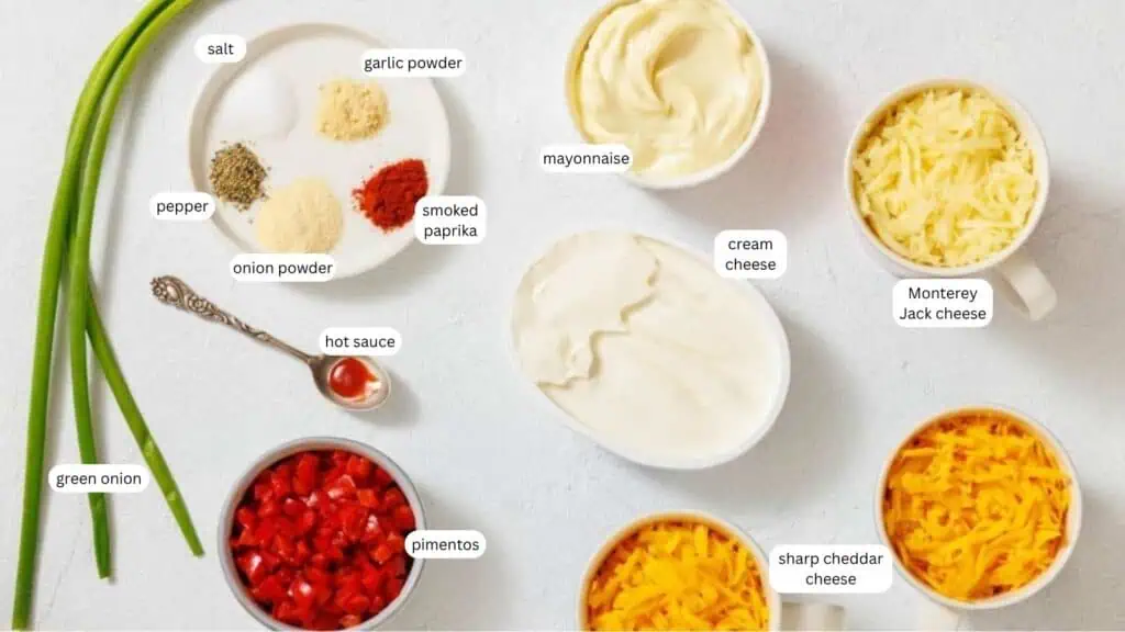 Various ingredients for a cheese spread, including green onion, pimentos, hot sauce, seasonings, mayonnaise, cream cheese, Monterey Jack cheese, and sharp cheddar cheese, arranged on a counter.