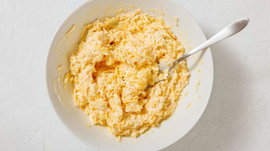 A white bowl filled with a creamy, shredded cheese mixture, with a metal fork resting inside, on a light-colored surface.