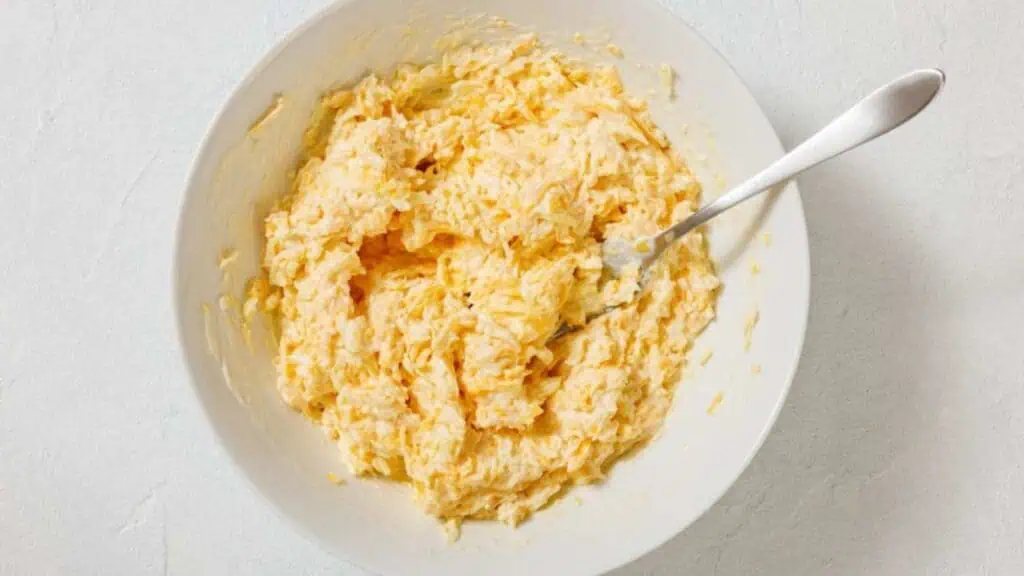 A white bowl filled with a creamy, shredded cheese mixture, with a metal fork resting inside, on a light-colored surface.