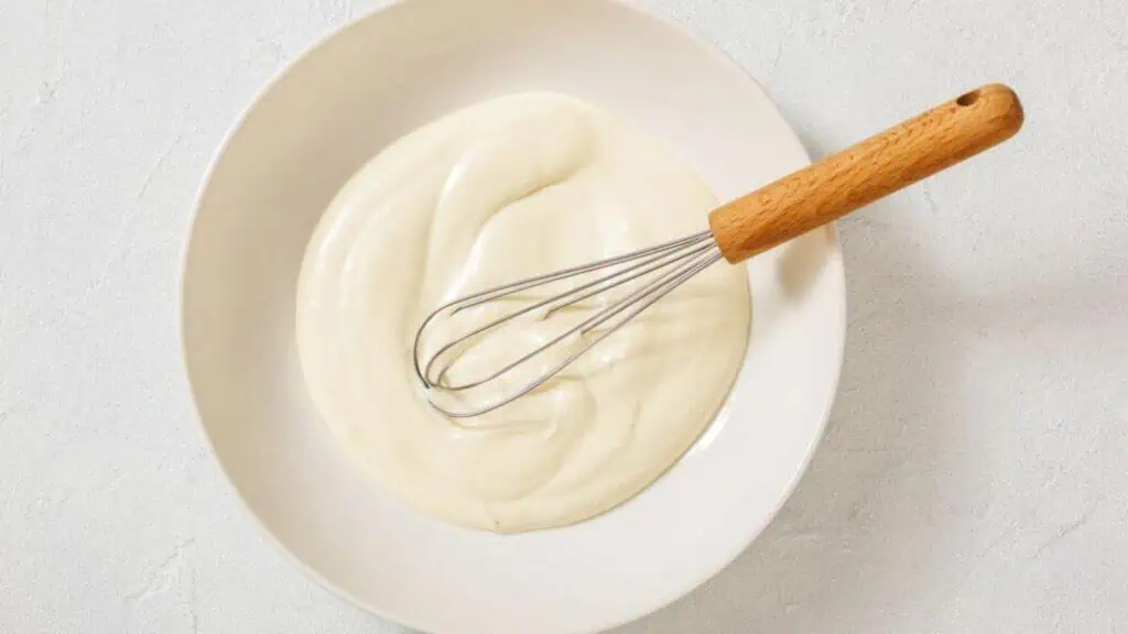 A metal whisk rests in a white bowl filled with a smooth, creamy white mixture on a light surface.
