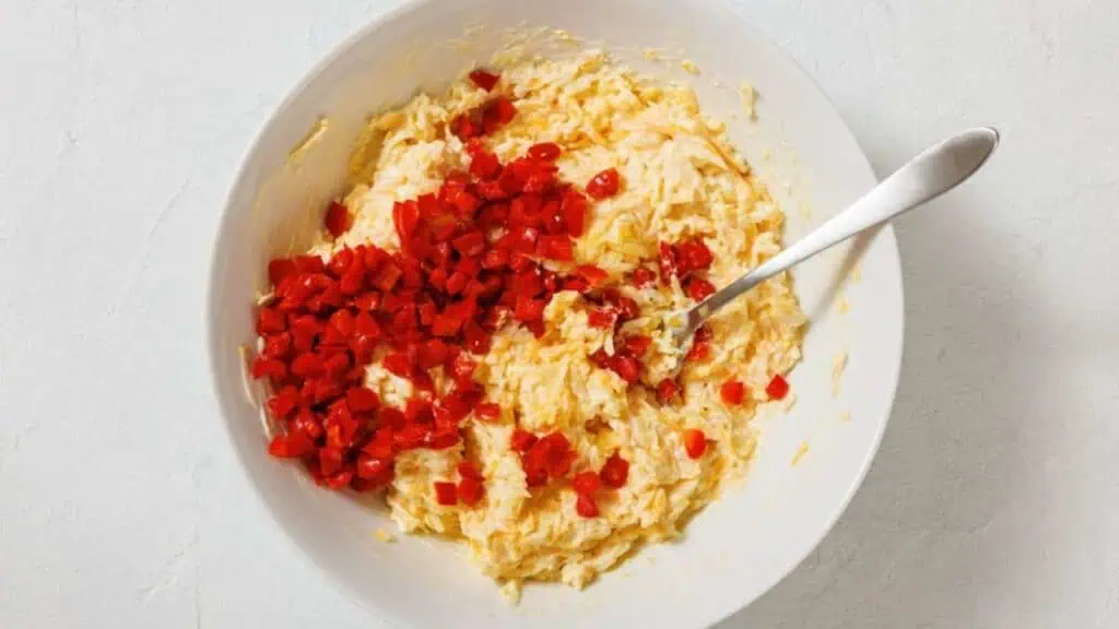 A bowl of shredded cheese mixture with chopped red pimentos on top and a spoon resting inside.