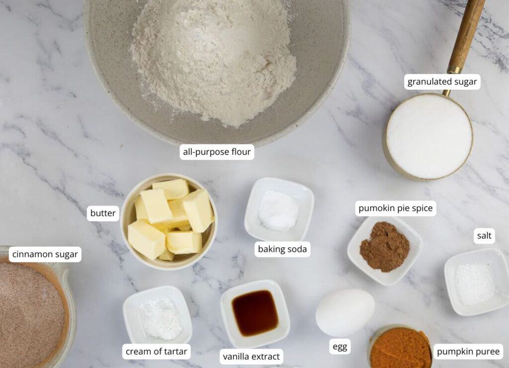 Baking ingredients measured in bowls on a marble counter, including flour, sugar, butter, spices, egg, pumpkin puree, cream of tartar, baking soda, salt, and vanilla extract.