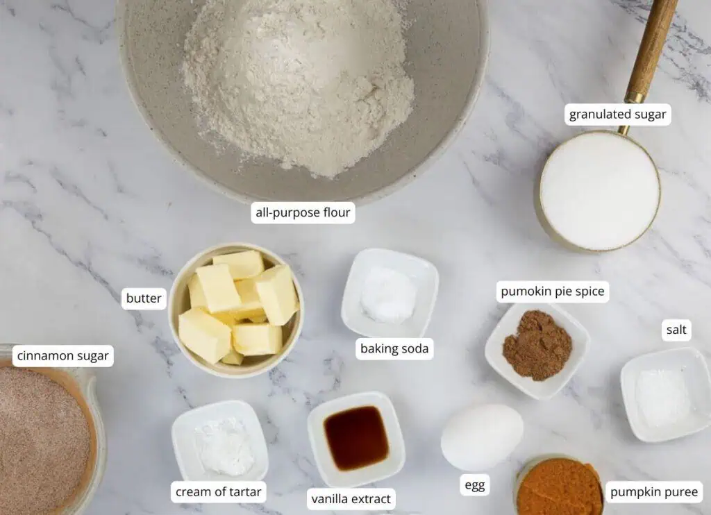 Baking ingredients measured in bowls on a marble counter, including flour, sugar, butter, spices, egg, pumpkin puree, cream of tartar, baking soda, salt, and vanilla extract.
