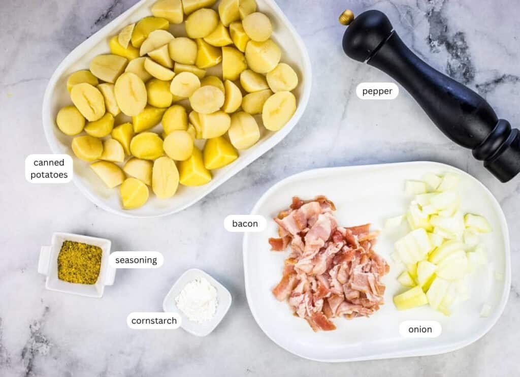 Sliced canned potatoes, chopped bacon, diced onion, seasoning, cornstarch, and a pepper grinder arranged on a countertop.