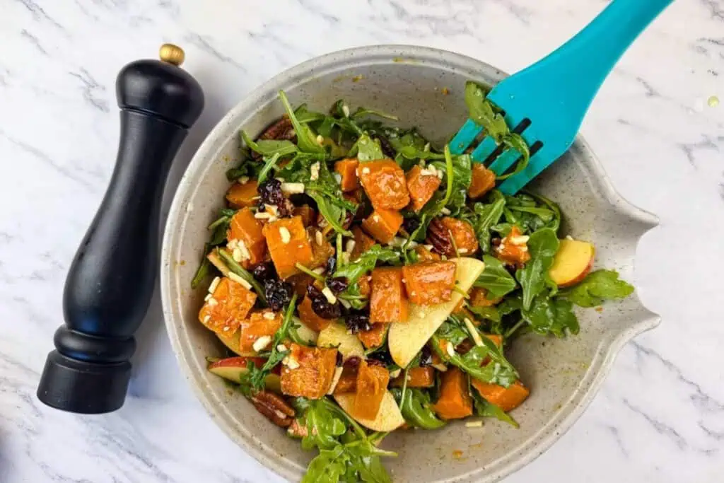 A bowl of salad with arugula, roasted sweet potatoes, apple slices, pecans, cheese, and dried cranberries, next to a black pepper grinder on a marble surface.