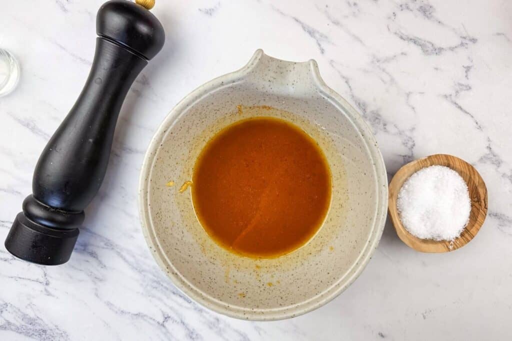 A bowl of brown liquid dressing sits on a marble surface next to a pepper grinder and a small wooden bowl of salt.