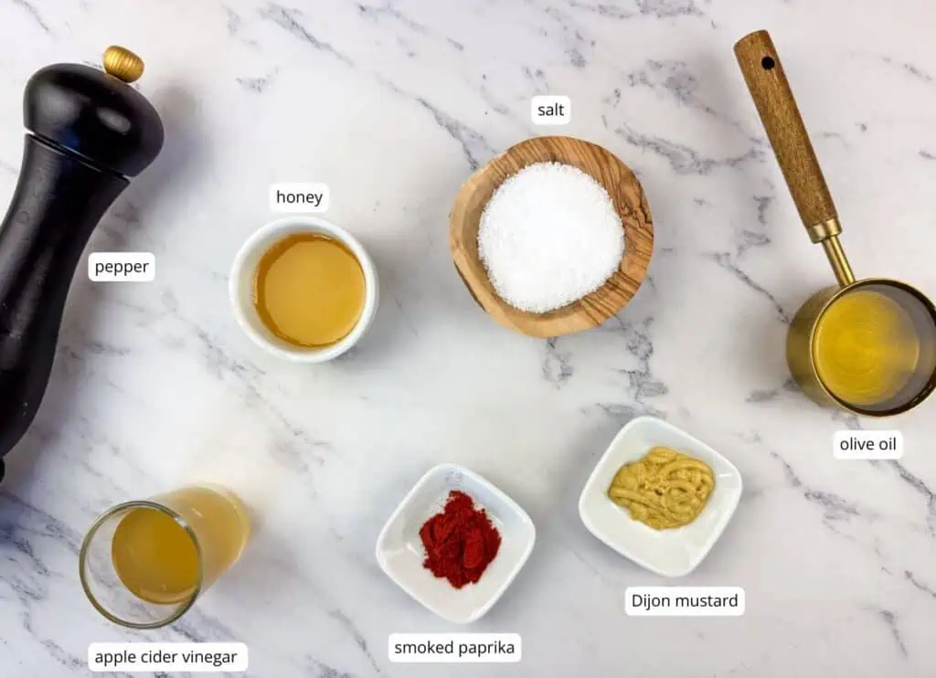 A marble surface displays labeled bowls of salt, pepper, honey, olive oil, apple cider vinegar, smoked paprika, and Dijon mustard.