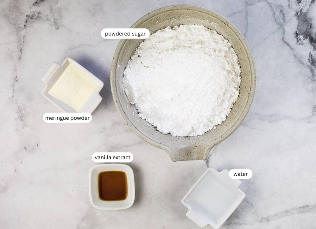 A bowl of powdered sugar, a small dish of meringue powder, a small dish of vanilla extract, and a small dish of water on a marble surface, each labeled.