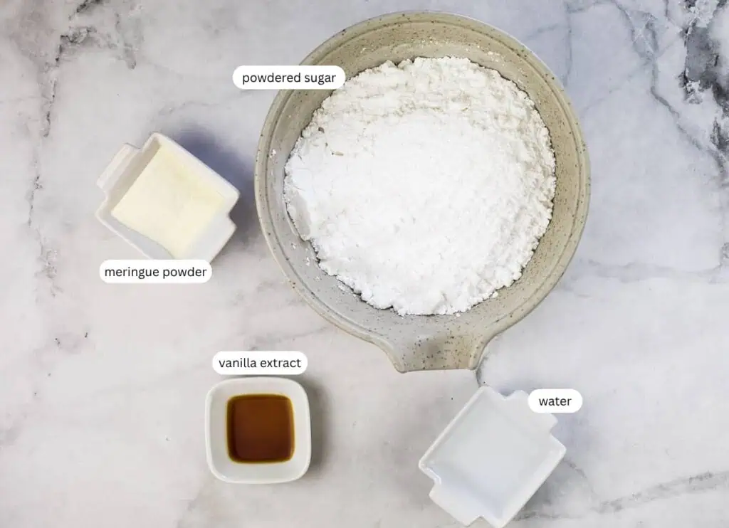 A bowl of powdered sugar, a small dish of meringue powder, a small dish of vanilla extract, and a small dish of water on a marble surface, each labeled.