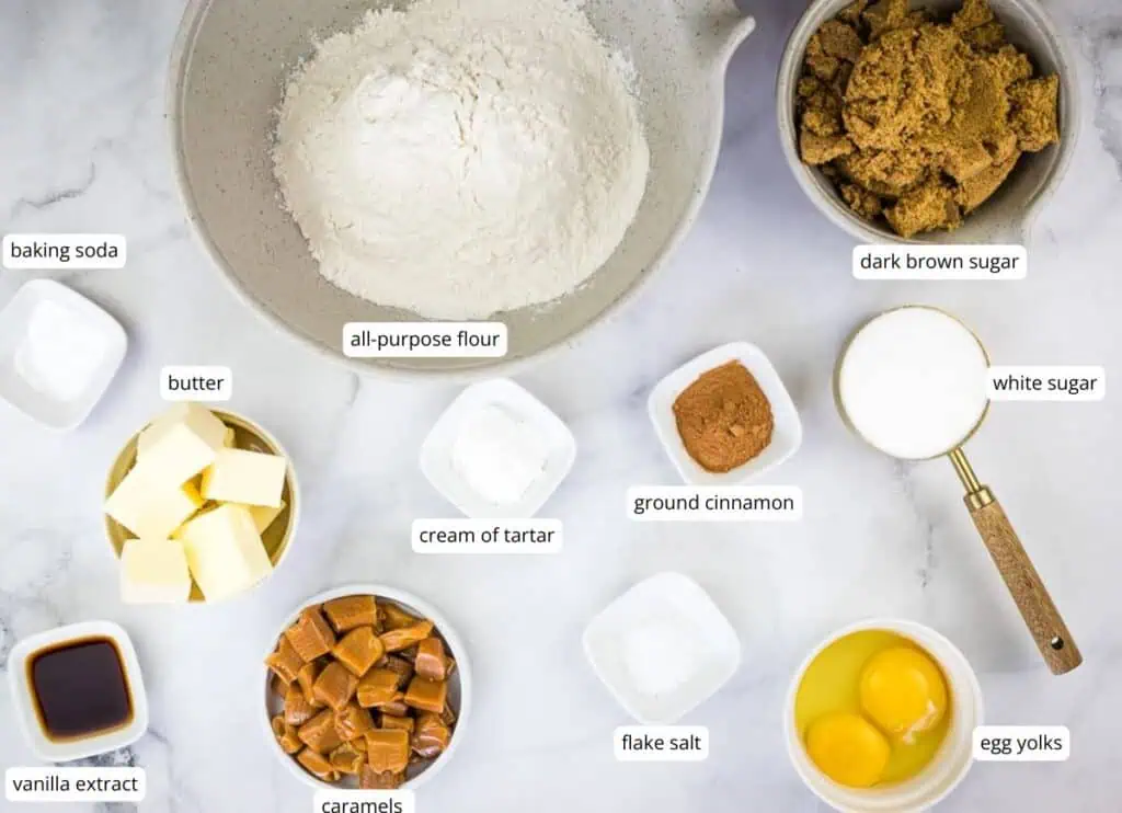 Baking ingredients on a white surface, including all-purpose flour, dark brown sugar, white sugar, butter, eggs, caramels, cinnamon, vanilla extract, baking soda, cream of tartar, and salt.