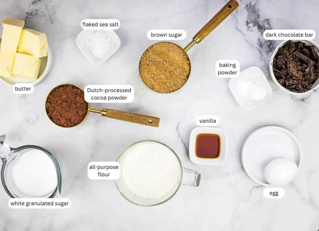 Various baking ingredients—such as butter, sugars, cocoa powder, flour, chocolate, egg, vanilla, baking powder, and flaked sea salt—are arranged on a marble surface.