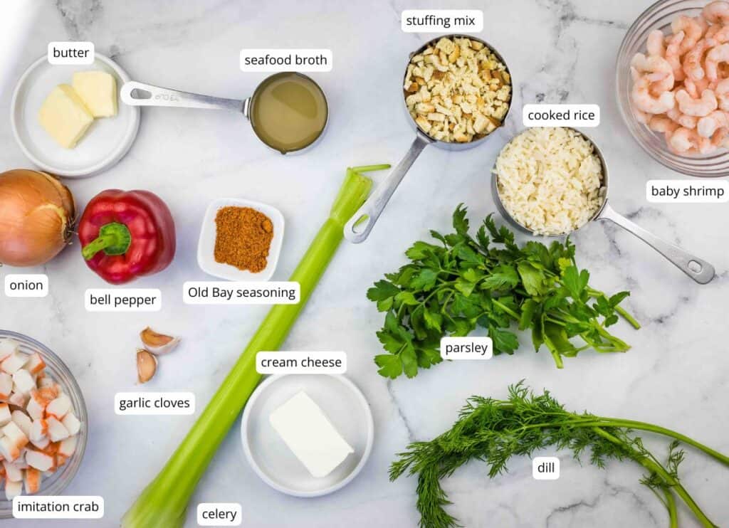 Ingredients for a seafood dish arranged on a white surface, including butter, onion, bell pepper, garlic cloves, imitation crab, celery, cream cheese, seafood broth, stuffing mix, cooked rice, baby shrimp, parsley, and dill.