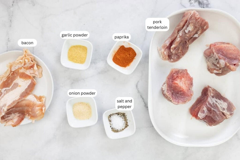 Raw pork tenderloin, bacon slices, and three small bowls of garlic powder, onion powder, and paprika, plus a small dish of salt and pepper on a white surface.