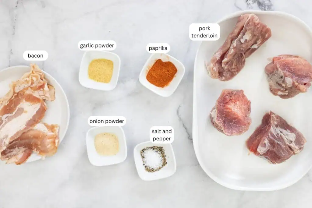 Raw pork tenderloin, bacon slices, and three small bowls of garlic powder, onion powder, and paprika, plus a small dish of salt and pepper on a white surface.
