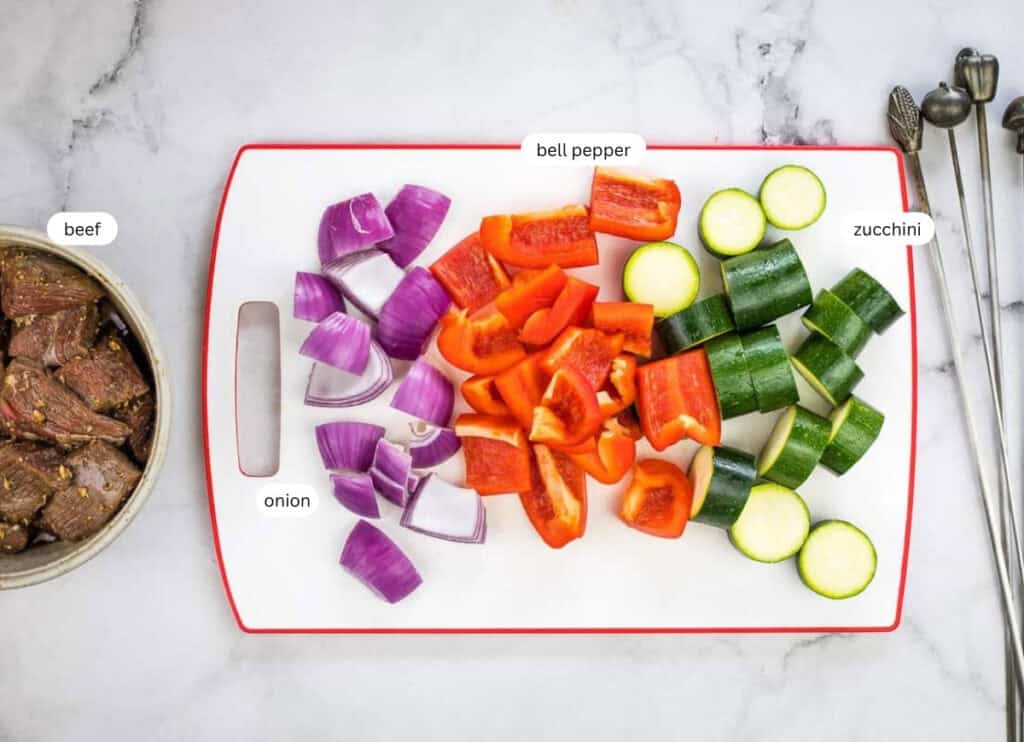 Chopped red onion, red bell pepper, and zucchini on a cutting board, with marinated beef pieces in a bowl nearby; ingredients labeled.