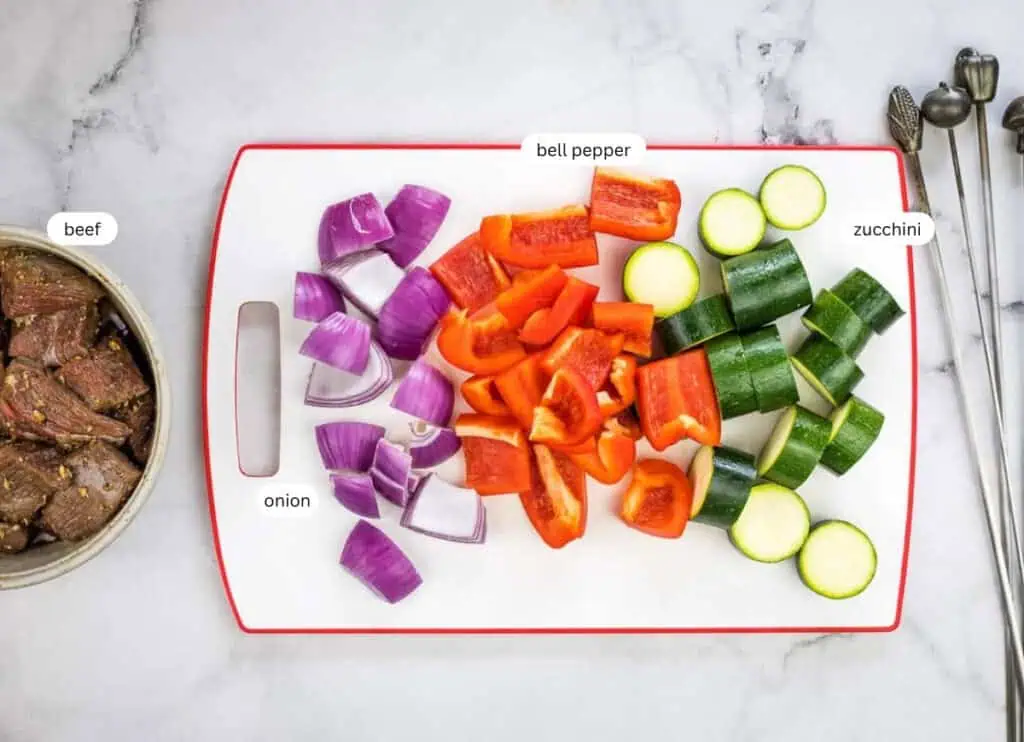 Chopped red onion, red bell pepper, and zucchini on a cutting board, with marinated beef pieces in a bowl nearby; ingredients labeled.