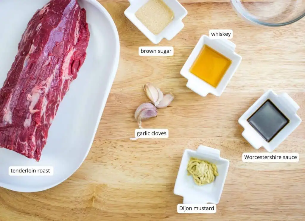 Top-down view of a tenderloin roast, garlic cloves, brown sugar, whiskey, Worcestershire sauce, and Dijon mustard on a wooden surface, each ingredient labeled.