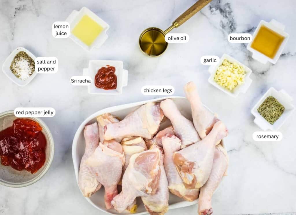 Raw chicken legs in a white dish surrounded by small bowls of labeled ingredients: lemon juice, olive oil, bourbon, garlic, rosemary, Sriracha, salt and pepper, and red pepper jelly.