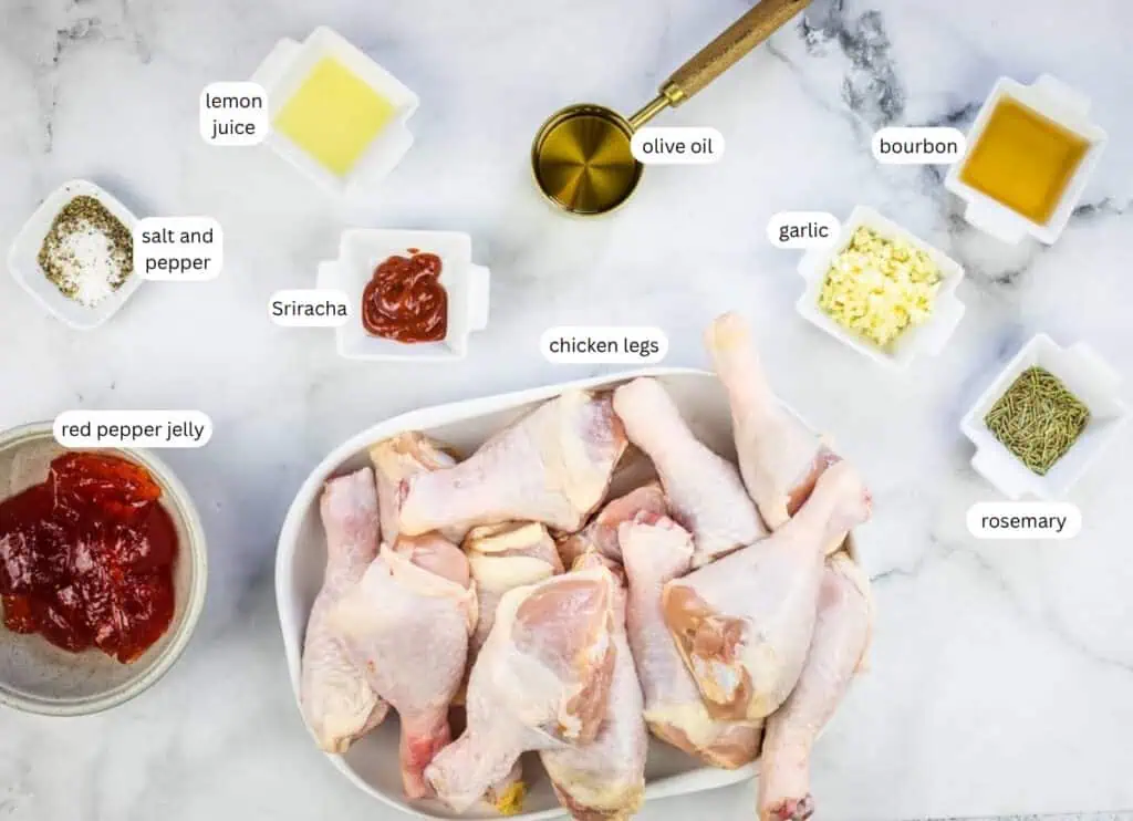 Raw chicken legs in a white dish surrounded by small bowls of labeled ingredients: lemon juice, olive oil, bourbon, garlic, rosemary, Sriracha, salt and pepper, and red pepper jelly.