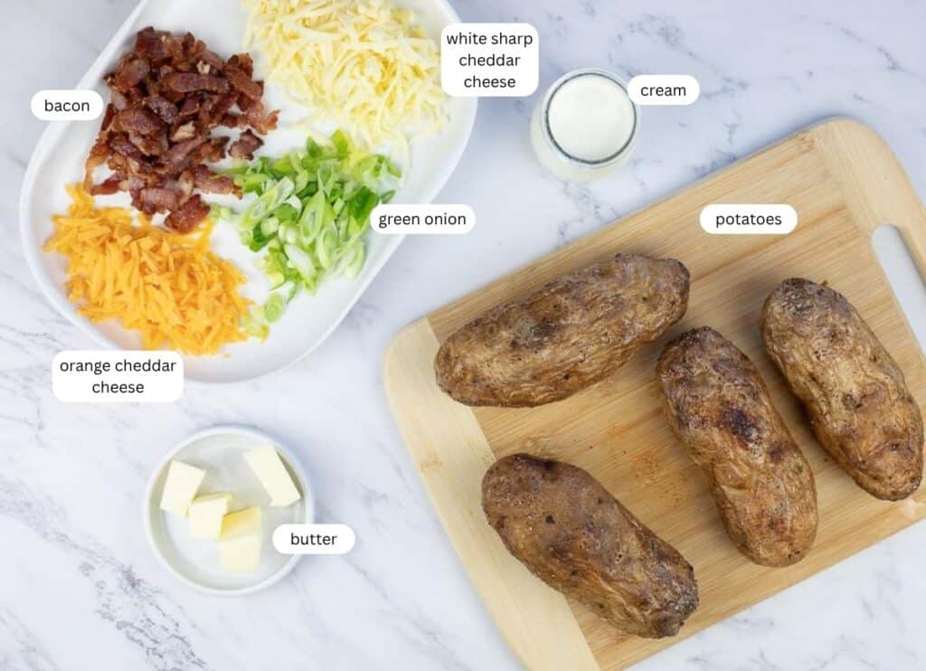 Ingredients for baked potatoes are arranged on a counter, including baked potatoes, chopped bacon, shredded cheddar cheeses, green onion, butter, and cream, all labeled.