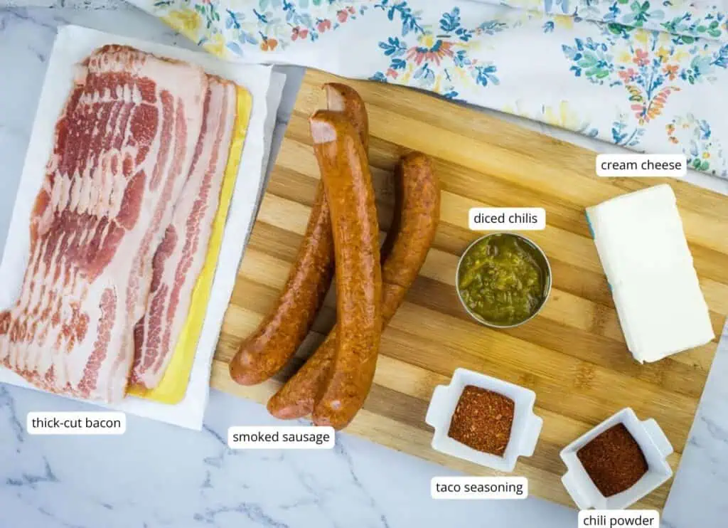 A wooden board with smoked sausage, diced chilis, cream cheese, taco seasoning, chili powder, and thick-cut bacon on a marble surface with a floral cloth.