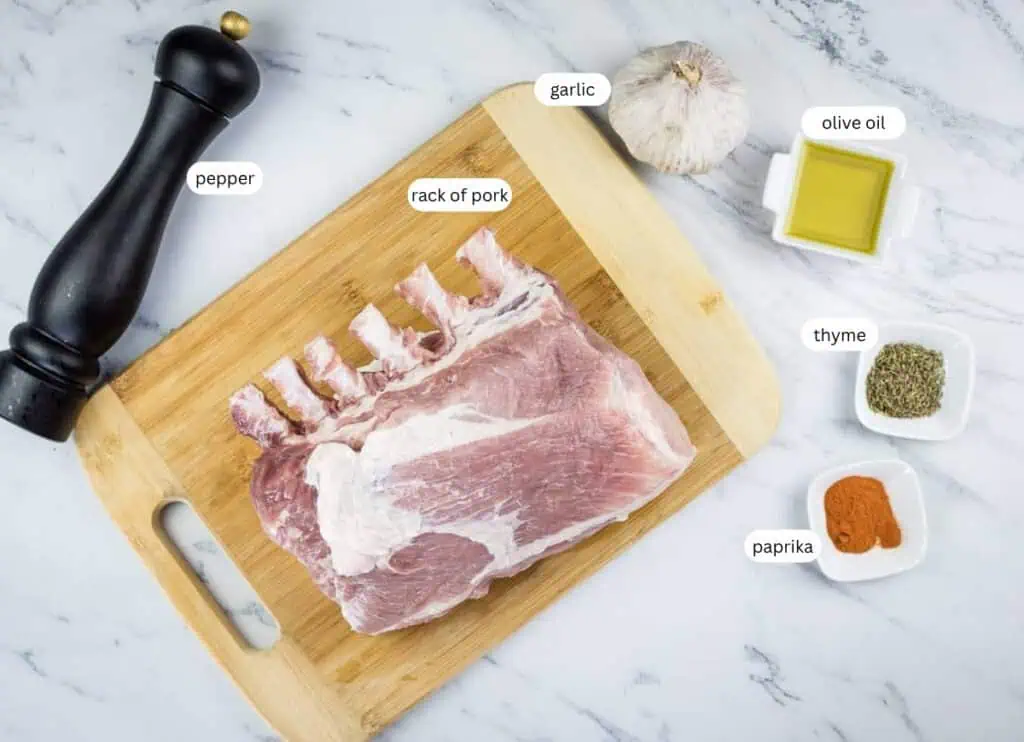 Raw rack of pork on a wooden cutting board with labeled ingredients: garlic, olive oil, thyme, paprika, and a pepper grinder on a marble countertop.