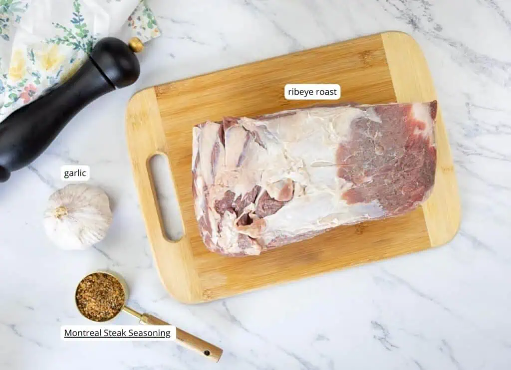 A raw ribeye roast on a wooden cutting board, with garlic, Montreal steak seasoning in a measuring cup, and a pepper grinder nearby, all on a marble surface.