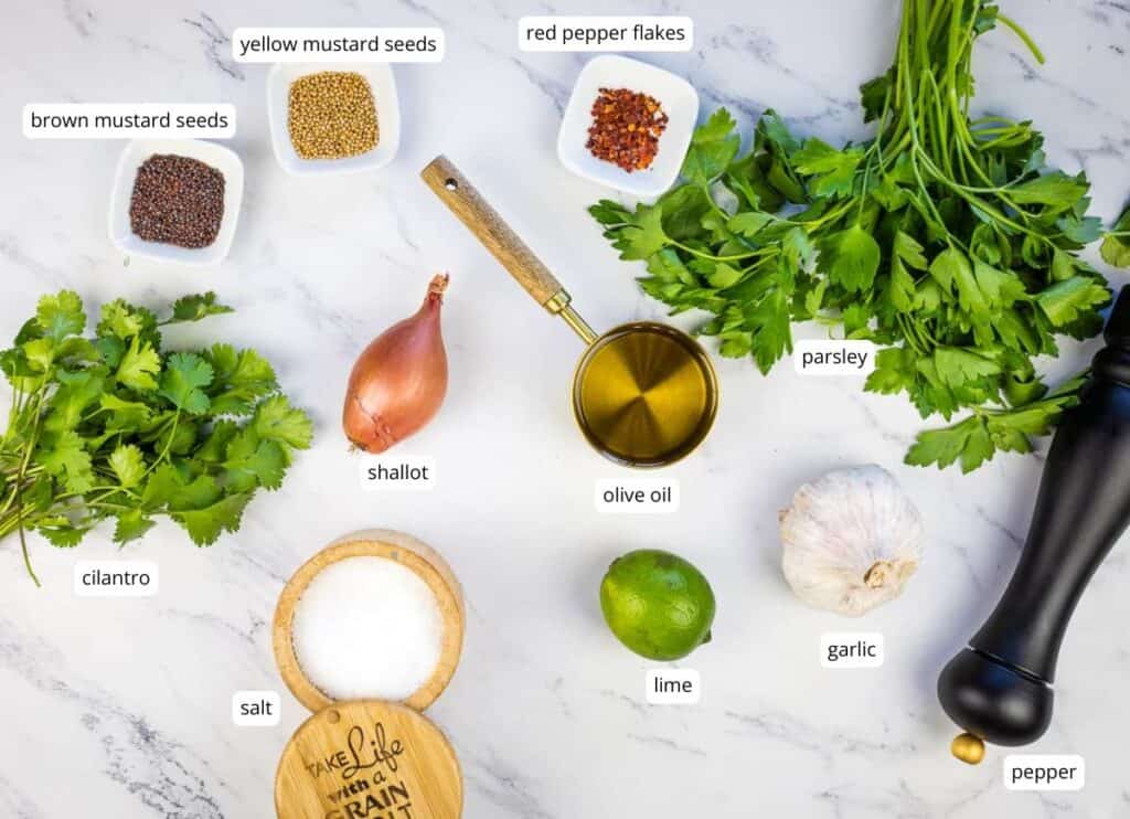 Ingredients on a white surface, including yellow and brown mustard seeds, red pepper flakes, parsley, cilantro, shallot, olive oil, lime, garlic, salt, and a pepper grinder.
