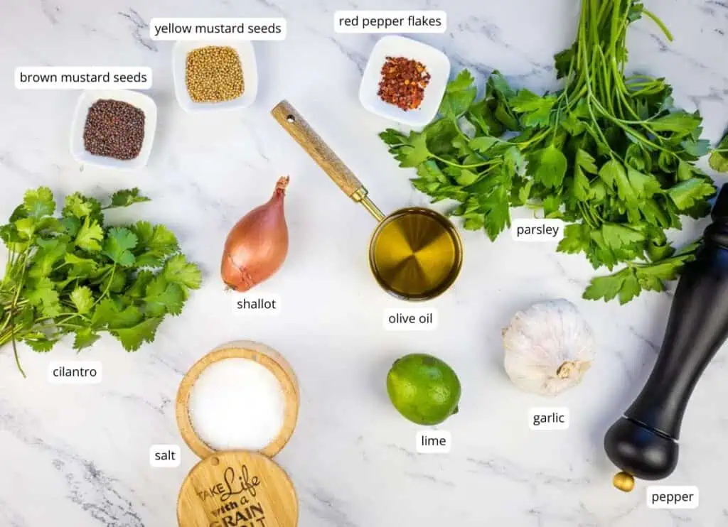 Ingredients on a white surface, including yellow and brown mustard seeds, red pepper flakes, parsley, cilantro, shallot, olive oil, lime, garlic, salt, and a pepper grinder.