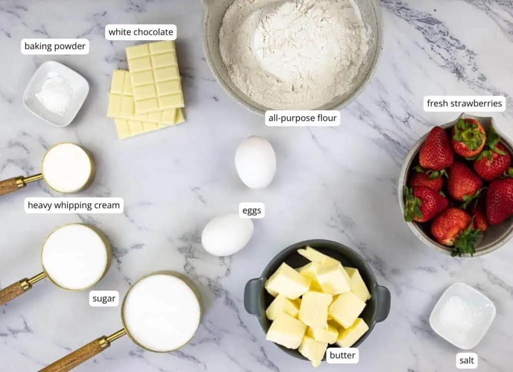 Top-down view of ingredients on a marble surface: white chocolate, all-purpose flour, fresh strawberries, eggs, butter, sugar, heavy whipping cream, baking powder, and salt.