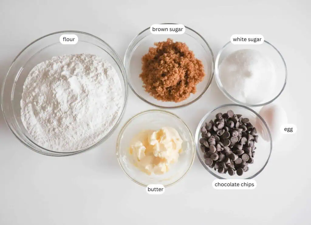 Five bowls containing labeled ingredients: flour, brown sugar, white sugar, butter, and chocolate chips, with a whole egg next to the chocolate chips.