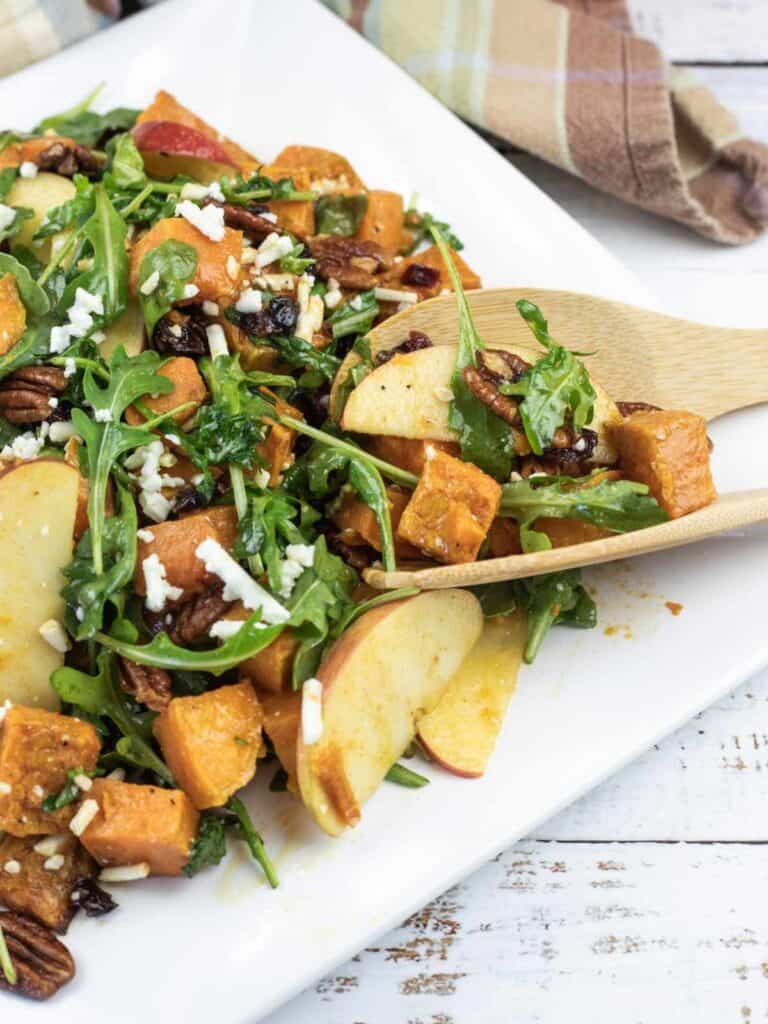 A salad with arugula, sliced apples, roasted sweet potatoes, pecans, cranberries, and crumbled cheese on a white plate with wooden utensils.