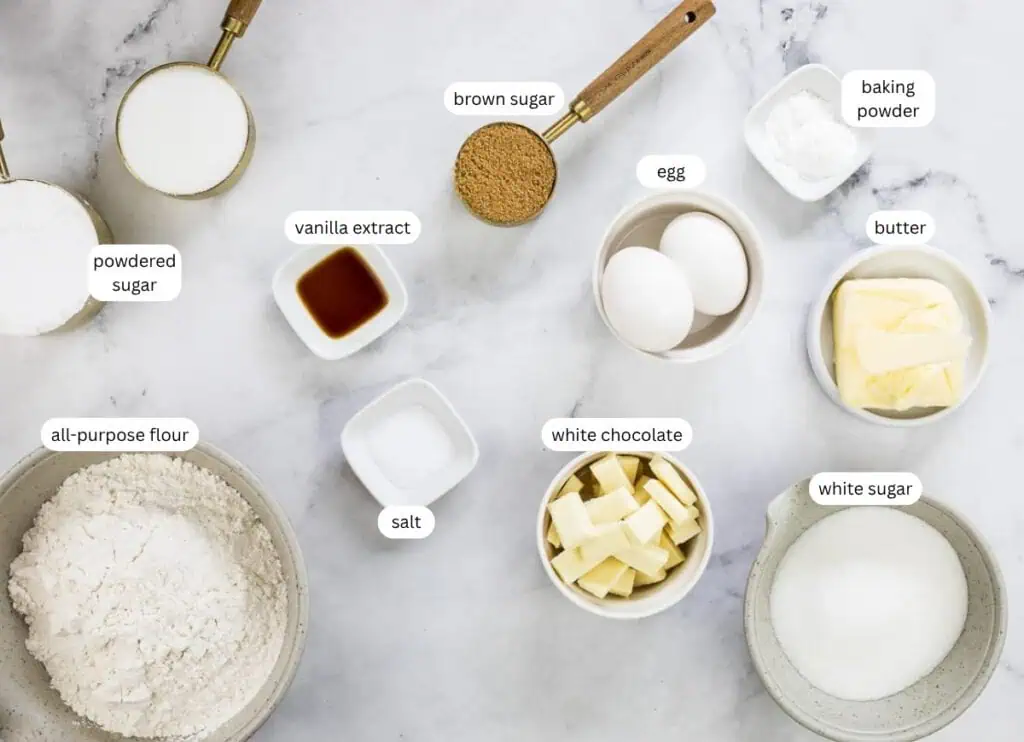 Baking ingredients arranged on a marble surface, including flour, powdered sugar, brown sugar, white sugar, salt, baking powder, butter, eggs, vanilla extract, and white chocolate.